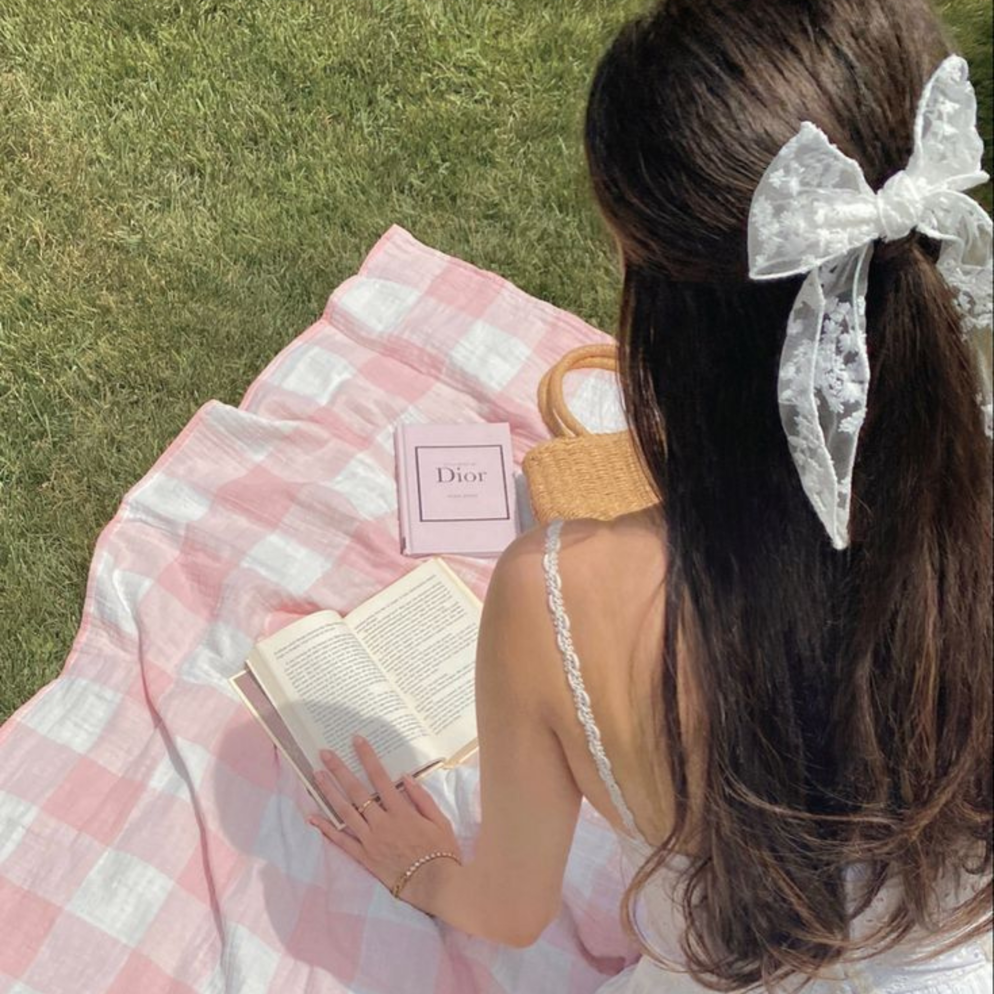 An image of a modern romantic woman sitting in a sundress on a picnic blanket in the grass reading a book, with a bow in her hair